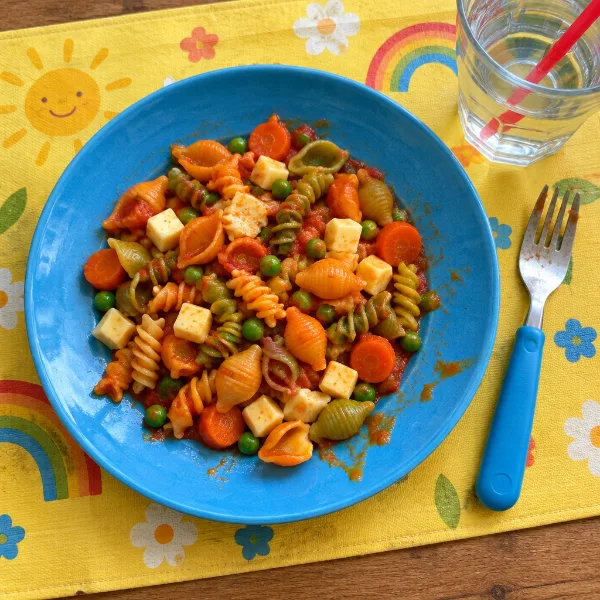 Kinderbord met bonte pastaschelpjes, tomatensaus, doperwten en worteltjes op een gele placemat